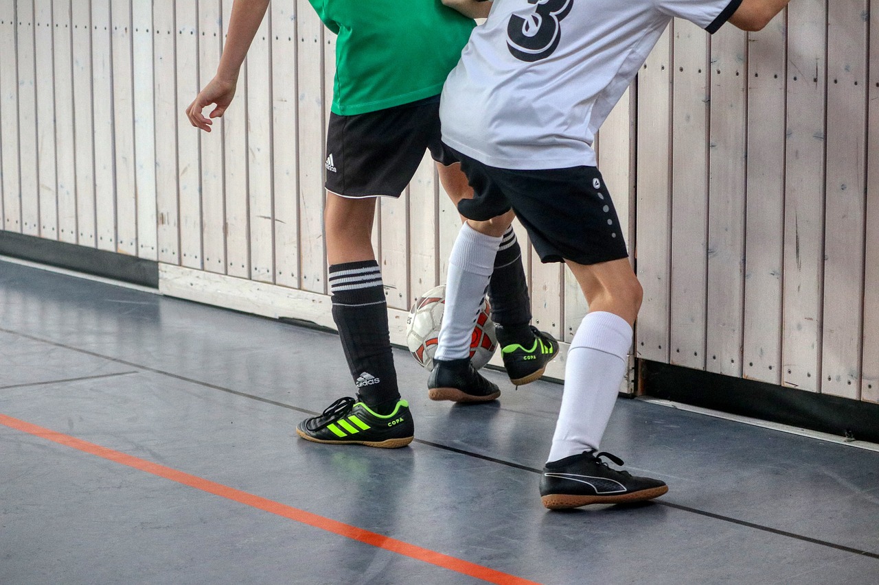 Zwei Hallenfußballspieler im Zweikampf nahe der Bande, mit schwarz-weißen Trikots und Sportschuhen auf dem Hallenboden.